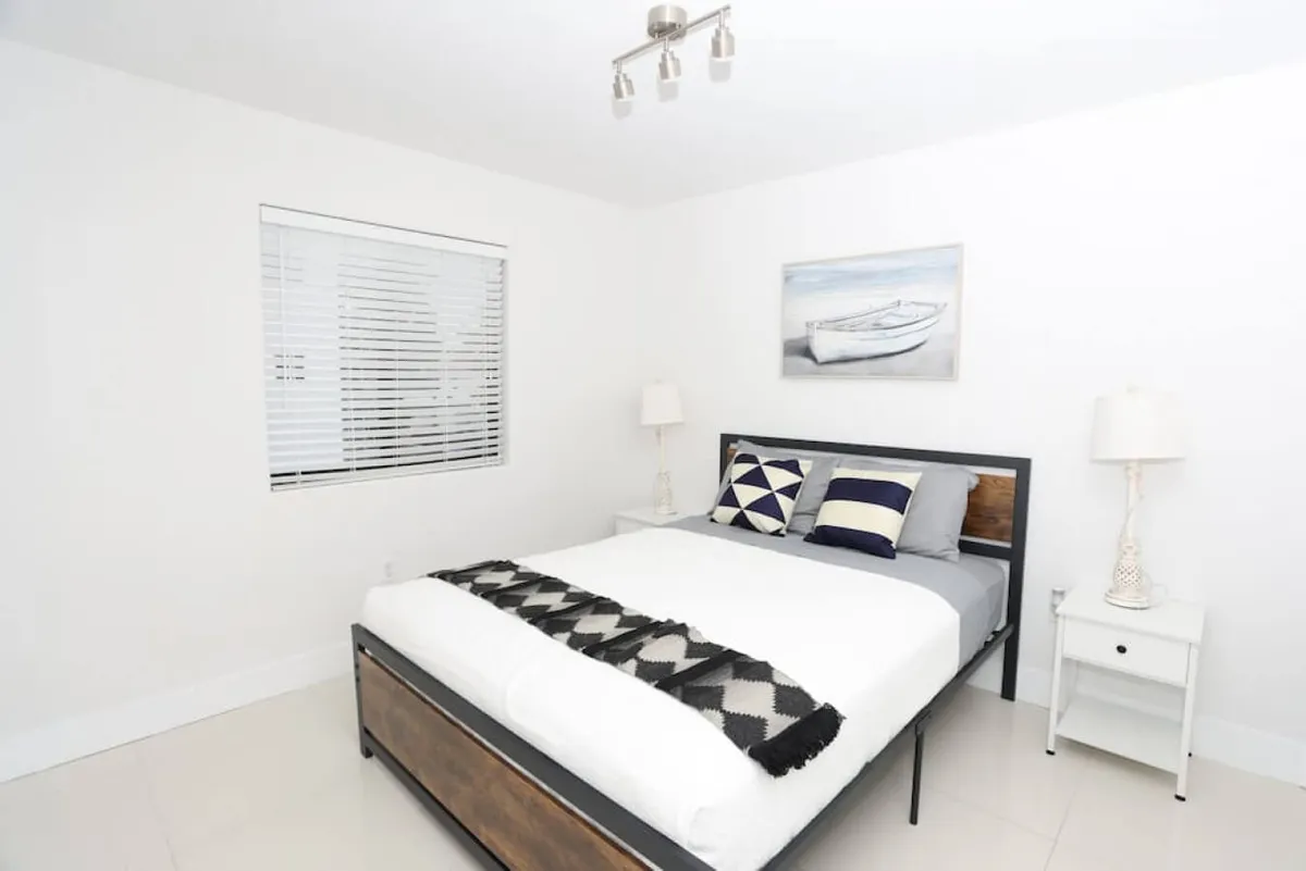 The image showcases a tranquil bedroom with a comfortable bed adorned with stylish geometric pillows, flanked by minimalist white bedside tables and warm lamps. Above the bed hangs a framed boat artwork, adding a touch of Miami flair, while natural light fills the uncluttered, inviting space.
