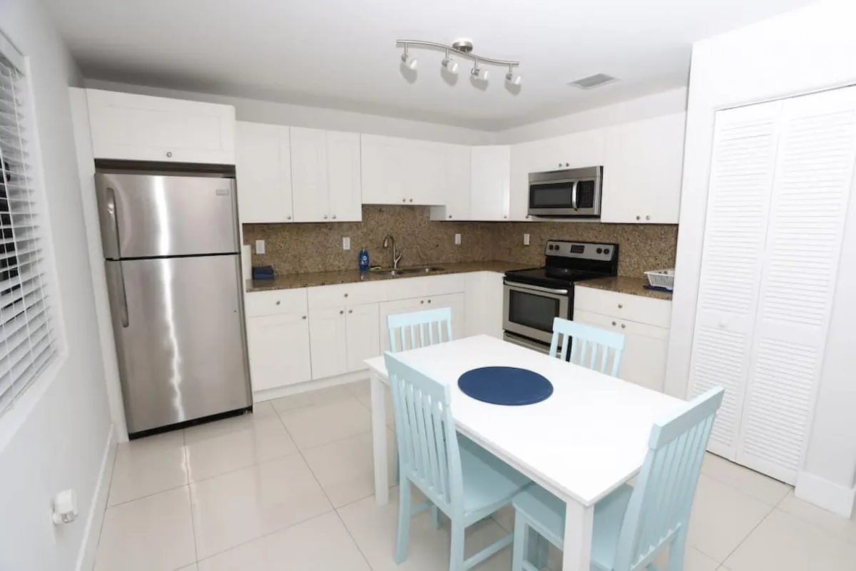 A bright and inviting kitchen featuring a stainless steel refrigerator, sleek white cabinetry topped with granite countertops, a modern stove and microwave, and a dining table with light blue chairs. The kitchen is open and thoughtfully arranged, creating a seamless flow between the cooking and dining areas.