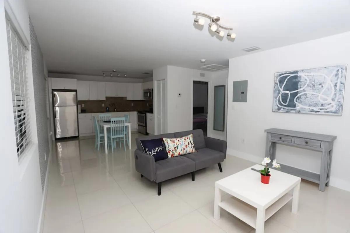 A thoughtfully arranged open-plan living and dining area combining a gray sofa with decorative pillows, a white coffee table with a small red plant, a light blue dining table with chairs, and a modern kitchen with stainless steel appliances. The overall ambiance is welcoming and comfortable, offering a space for relaxing, dining, and socializing.
