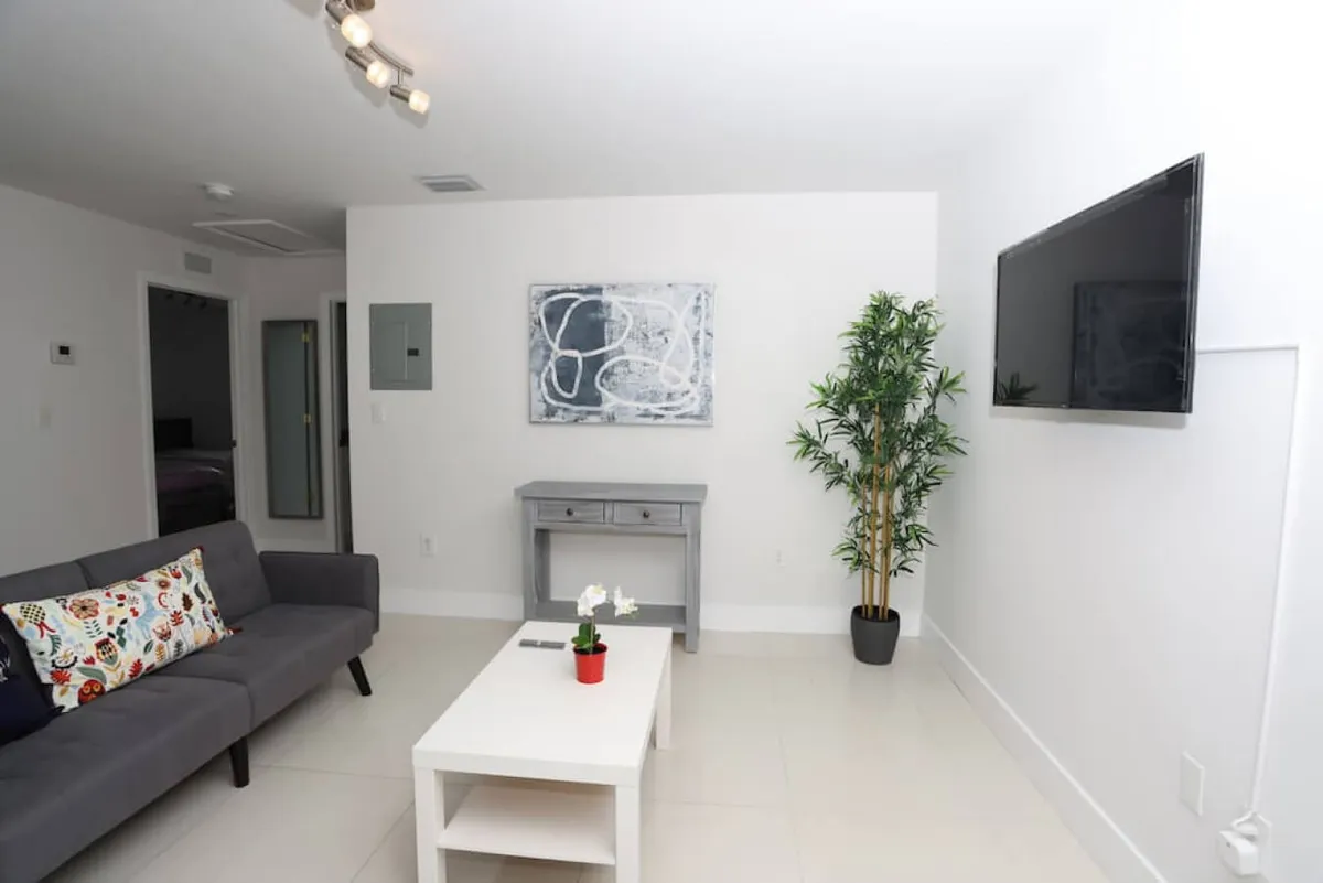 A view of the living room featuring a plush grey sofa adorned with a colorful patterned pillow, a white coffee table with a small red pot plant, a flat-screen TV mounted on the wall, and a tall green decorative plant in the corner. Natural light pours in, creating a calm and inviting atmosphere ideal for both relaxation and socializing.