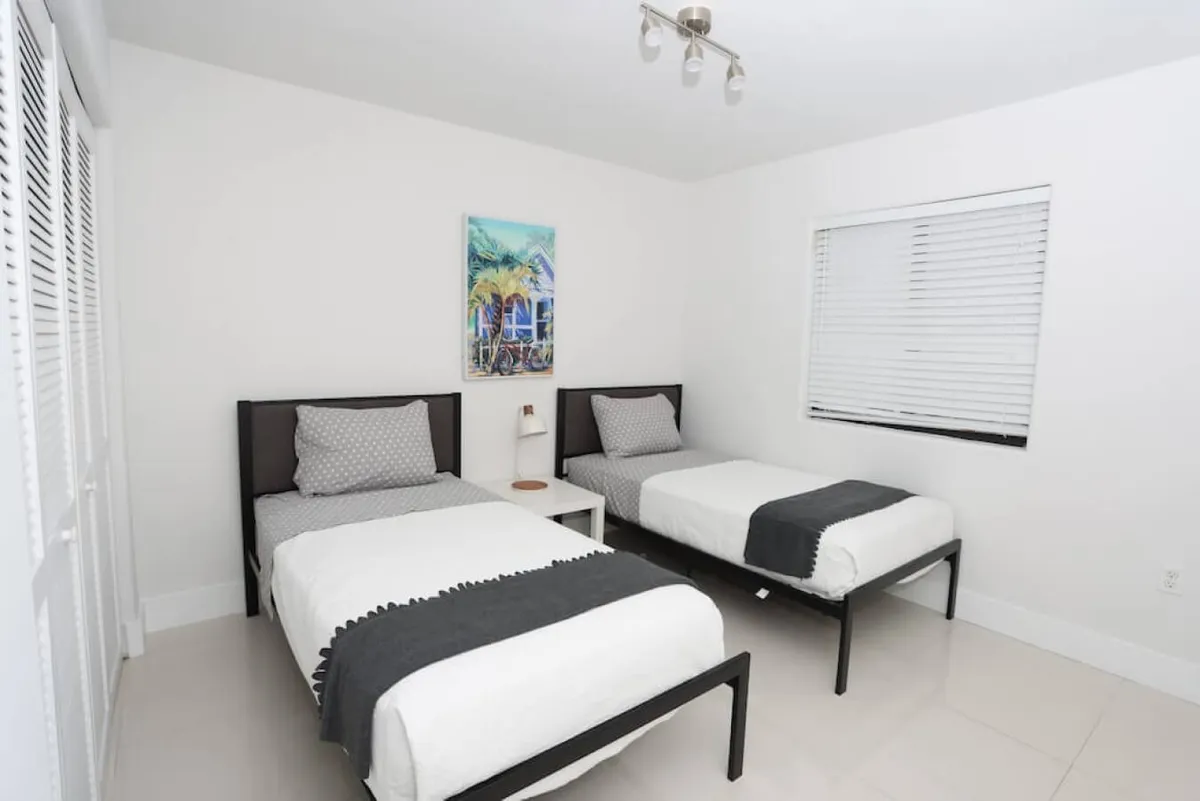 This image shows a tranquil bedroom with two single beds that feature dark frames and matching bedding. A small white nightstand sits between the beds, topped with a lamp, while colorful wall art depicting a tropical scene hangs above. Natural light filters through the window with white blinds, enhancing the room's restful ambiance.