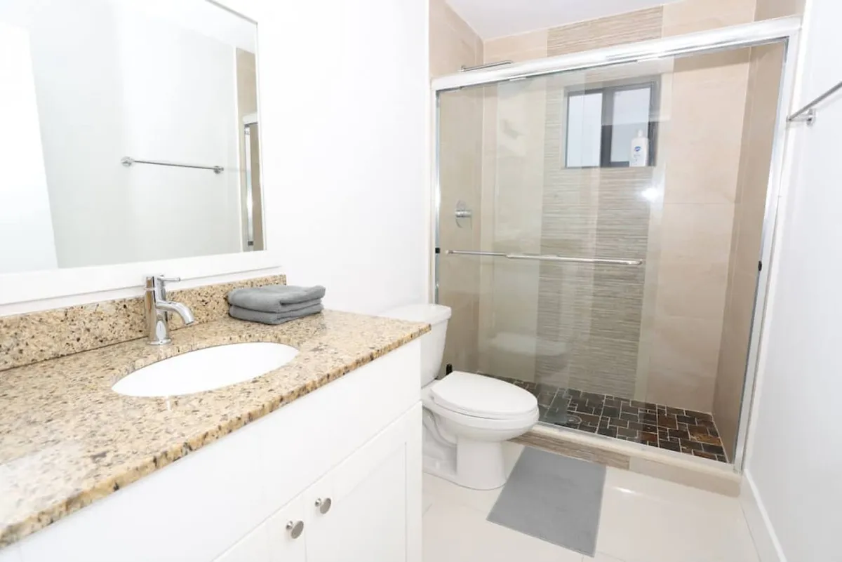 This bathroom features a modern granite countertop with a sink, a sparkling clean toilet, and a glass-enclosed shower with a tiled wall. The space is bright and fresh, emphasizing cleanliness and comfort.