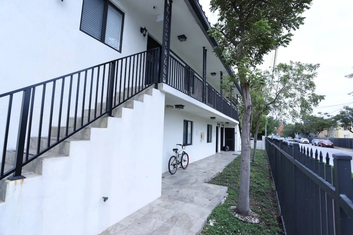 This photo captures the exterior staircase leading up to the second-floor apartment entry, with a black metal railing and a bicycle parked near the entrance. Lush greenery from a nearby tree adds to the peaceful ambiance, and the image hints at the secure, elevated entryway.