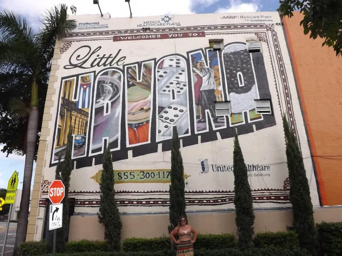 A vibrant street scene featuring a large mural with 'Little Havana' text, filled with colorful cultural imagery inside each letter. Street signs and a pedestrian crossing add an urban context, while a person poses in front of the mural, capturing the moment and the essence of the neighborhood.