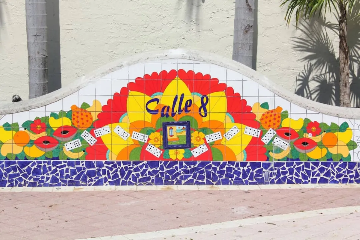 An exterior view of a vibrant mosaic mural emblazoned with 'Calle 8,' adorned with bright floral and fruit motifs and playful domino tiles. Palm trees cast dappled shadows, enhancing the festive, tropical vibe. The scene suggests socializing and exploration along this iconic street.